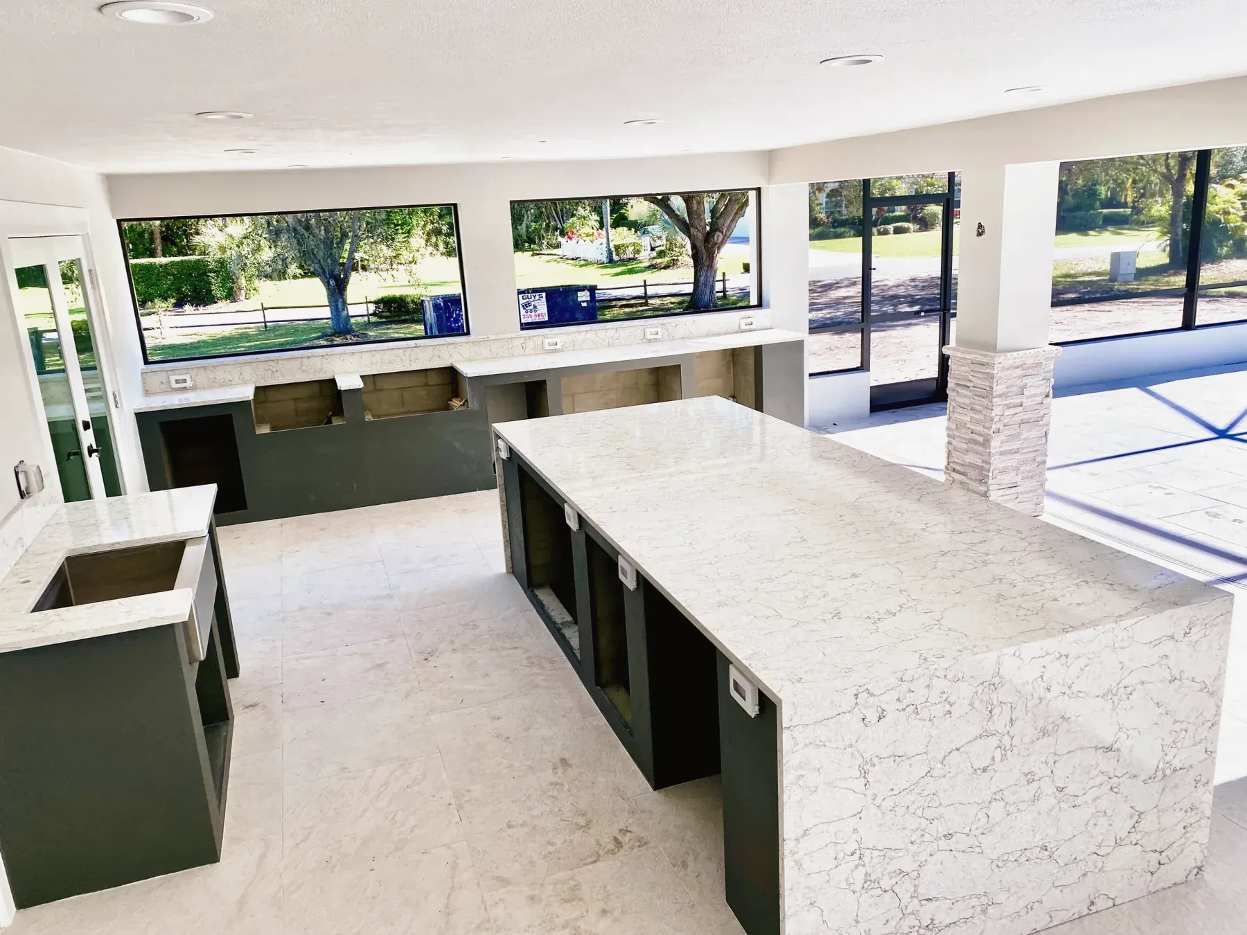 Covered lanai with a long white veined waterfall-edge island, stacked-stone columns and large windows