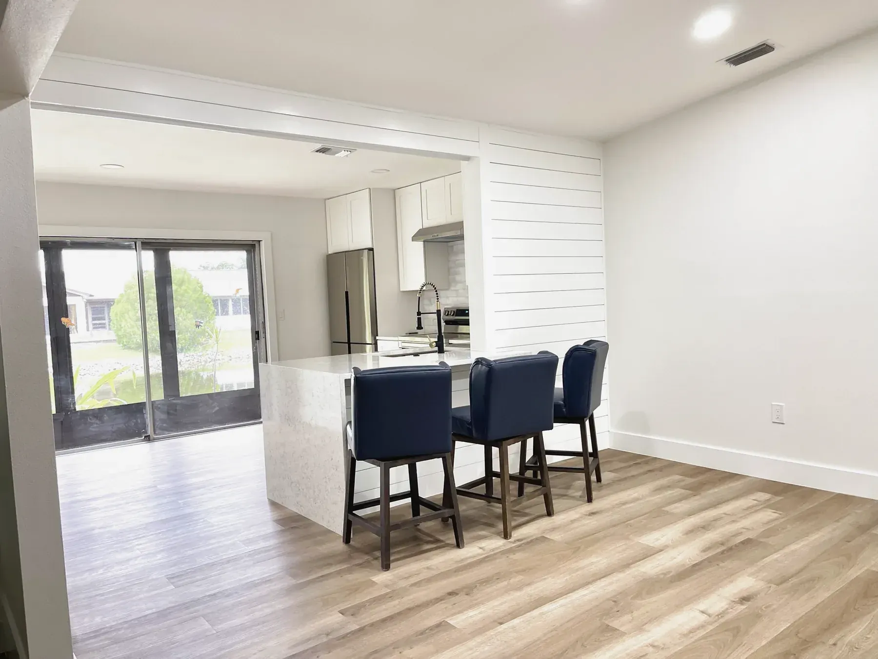 Open kitchen with a white waterfall-edge peninsula, three blue bar stools and a shiplap wall