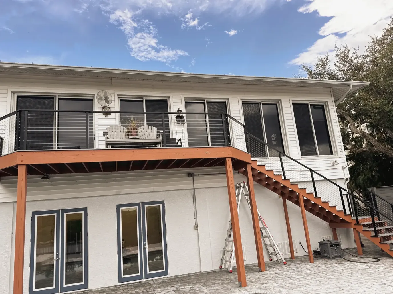 White two-story house with an elevated deck, black railings and a staircase above a paver patio
